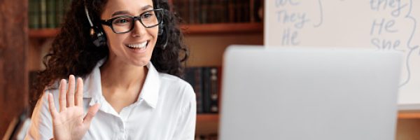 Distance Education. Positive lady wearing glasses and wireless headest at virtual meeting, sitting at desk, having video call on laptop, waving to webcam. Woman studying or teaching online at home